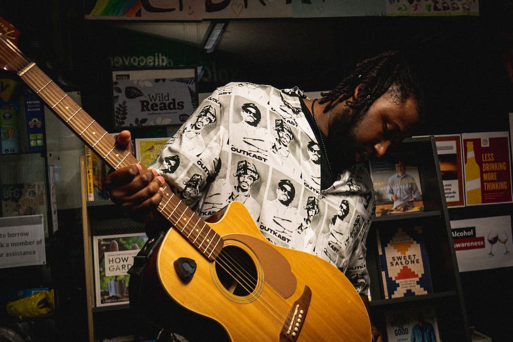 Bookmarks and Basslines tile image resize A musician playing a guitar in a library space. Credit: Luey Northern