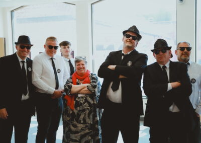 Jo, manager of Lowestoft Library, posing alongside a group of men wearing black suits, sunglasses and fedoras.